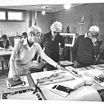 Group of artists in a classroom setting reviewing and discussing artwork on tables.