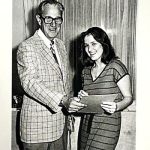Older man presenting a certificate or award to a young woman in a striped dress.