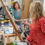 Two women discussing artwork in busy, creative studio environment.