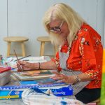 Woman painting at table, focused and smiling in floral shirt.