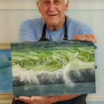 Man proudly holding realistic wave painting in art classroom.
