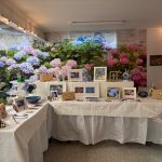 Table display of floral artwork and crafts with hydrangea backdrop.