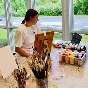 Woman painting in studio surrounded by brushes and paint bottles